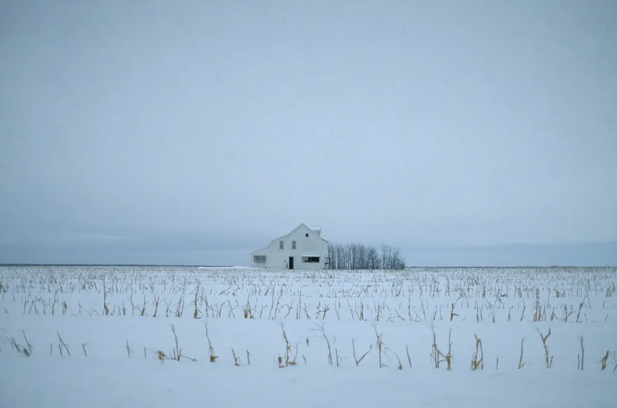 A white Iowa farmhouse alone in a snow-covered field of corn stubble under a pale winter sky